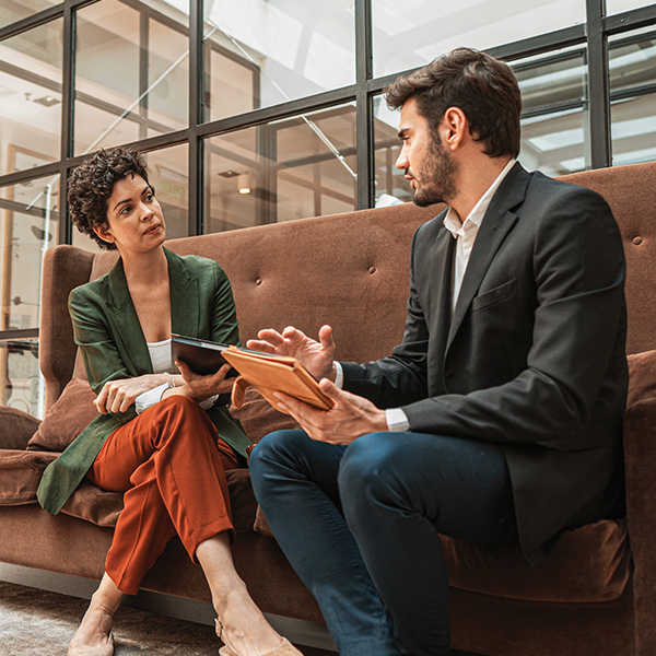 Two people dressed in business attire seated on a sofa, engaged in conversation.